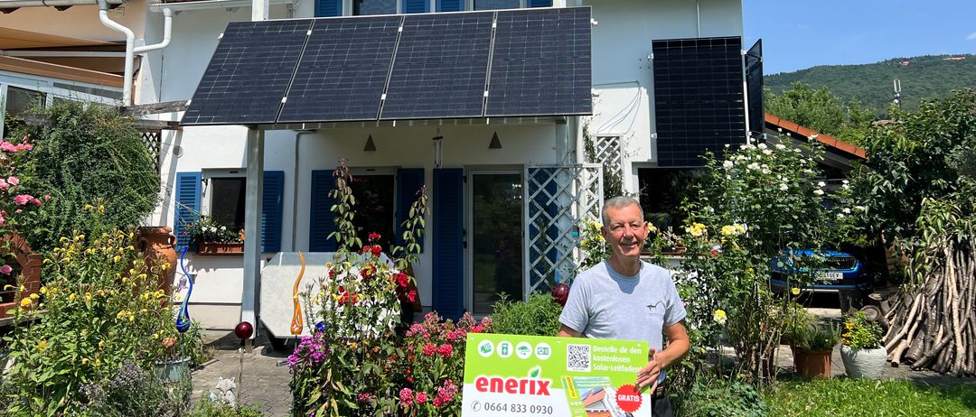 Ein Mann steht vor einem Haus mit Solarpanelen auf dem Dach und im Garten. Er hält ein grünes Banner mit Text und einem QR-Code.