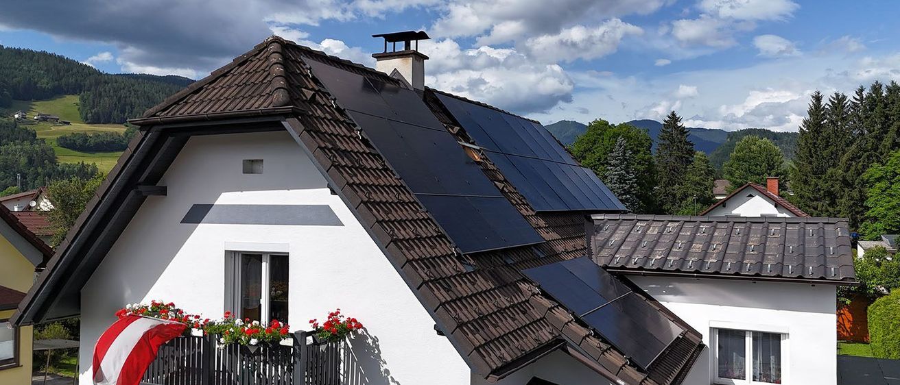 Ein weißes Haus mit einem braunen Dach und Solarmodulen, einem Balkon mit roten Blumen und einem Schornstein unter einem teilweise bewölkten Himmel.