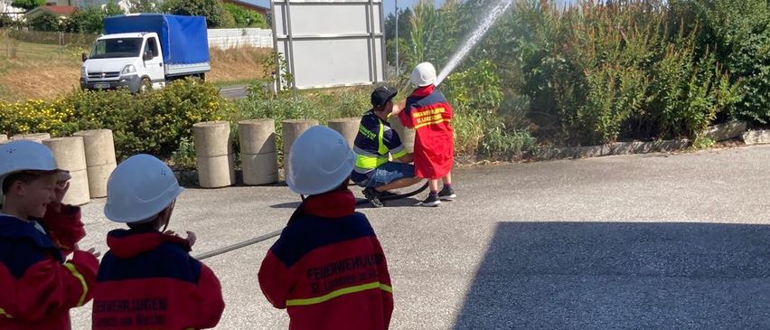 Drei Kinder in roten Uniformen beobachten einen Feuerwehrmann, der einen Schlauch auf einem Grasbereich mit Büschen und einem Lastwagen im Hintergrund einsetzt.