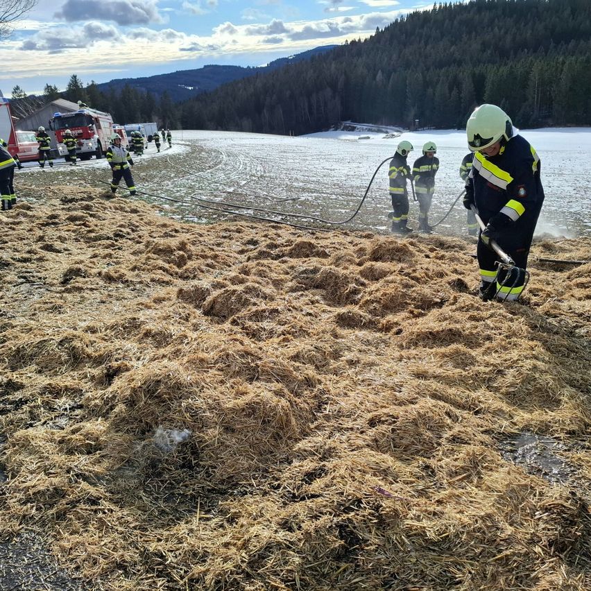 Eine Gruppe von Feuerwehrleuten arbeitet auf einem verschneiten Feld, rückt mit Heu und Ausrüstung um. Dahinter sind Feuerwehrwagen und ein Gebäude zu sehen.