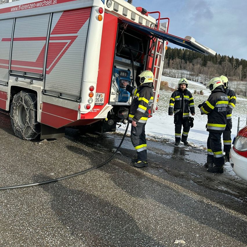 Drei Feuerwehrleute stehen auf einer verschneiten Straße, einer benutzt einen Schlauch von einem roten LKW. Sie tragen alle Helme und Handschuhe. Die Straße ist nass, und ein weißes Auto ist in der Nähe.