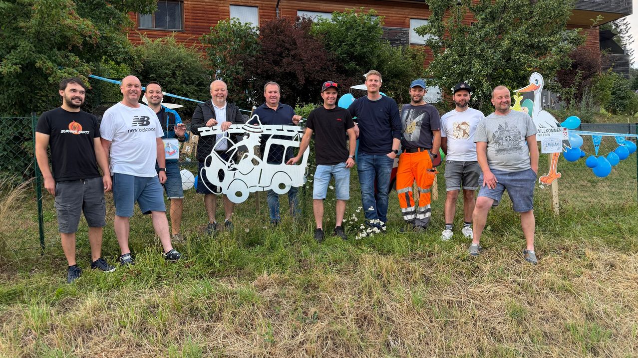 Eine Gruppe von Männern steht auf einem Feld mit einer großen weißen Ausschnitt eines Busses. Sie lächeln und scheinen für ein Foto zu posieren. Hinter ihnen sind Bäume und ein Gebäude mit Fenstern.