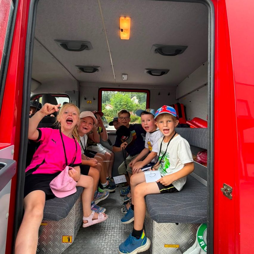 A group of children are sitting inside a red fire truck, smiling and posing for a photo. The interior is equipped with seats and storage compartments.