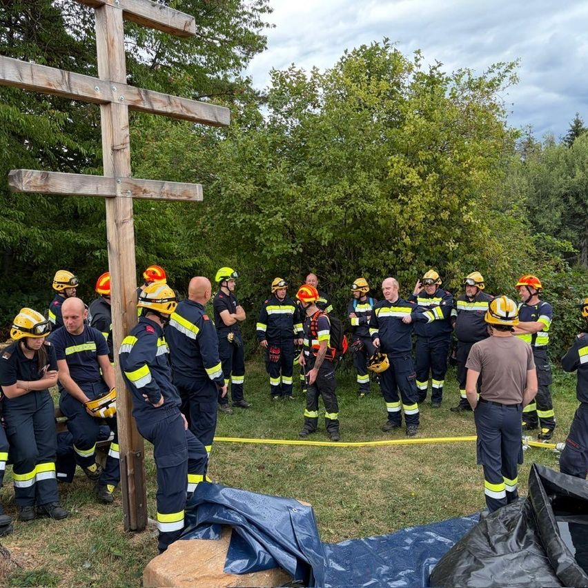 A group of firefighters in uniform, some with helmets, are gathered around a wooden cross in a grassy area with trees and cloudy sky.