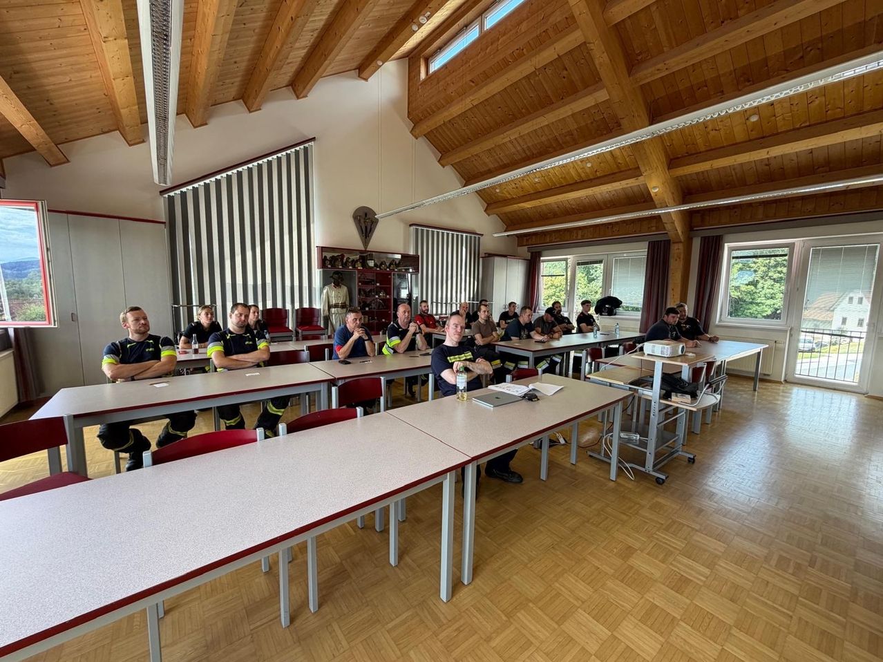 A group of firefighters in uniform are sitting at tables in a classroom. The room has wooden beams and large windows.