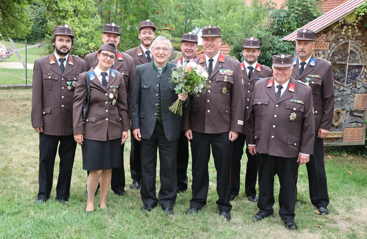 Eine Gruppe uniformierter Personen, darunter ein Mann mit Brille und eine Frau, stehen in einem Garten zusammen, wobei ein Mann einen Blumenstrauß hält.