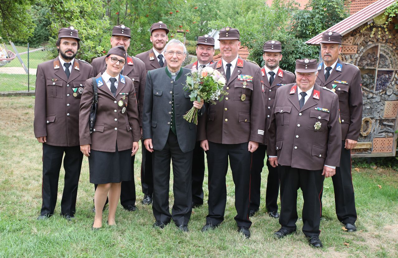 A group of uniformed individuals, including a man in glasses and a woman, stand together in a garden with a man holding a bouquet of flowers.