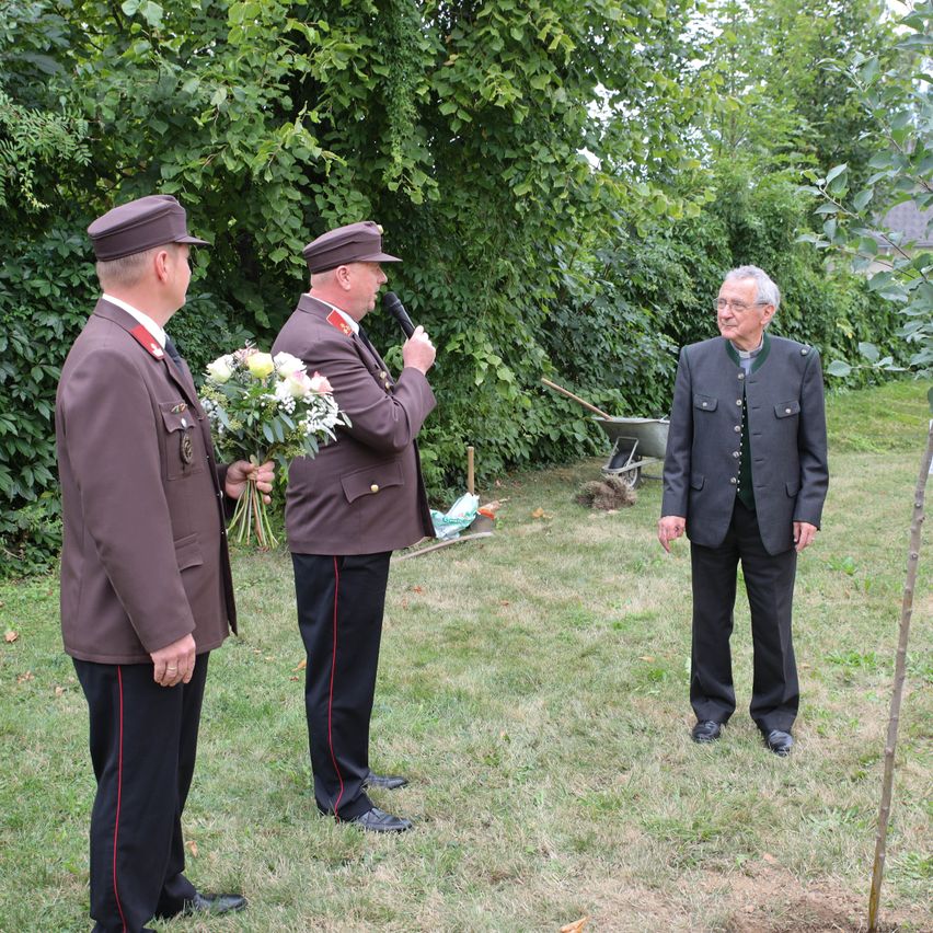 Three men in uniform stand in a garden, one holding a bouquet of flowers and a microphone, while another man speaks into the microphone.