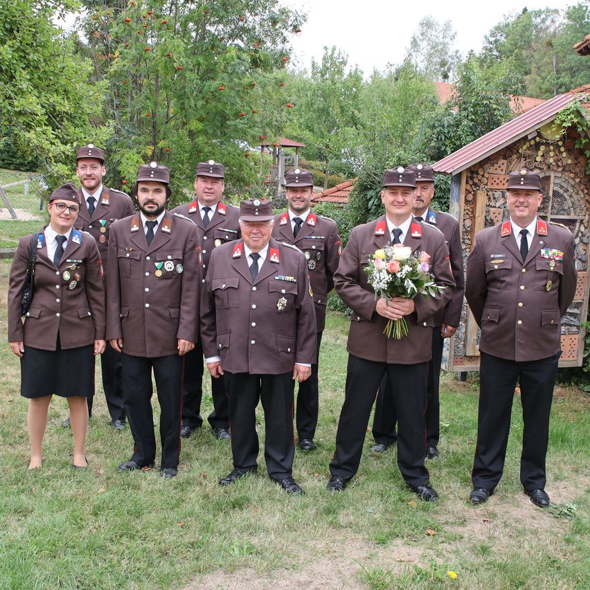 A group of uniformed individuals, possibly a military or police unit, stands in a line with one person holding a bouquet of flowers. They are in front of a small wooden house with a red roof.