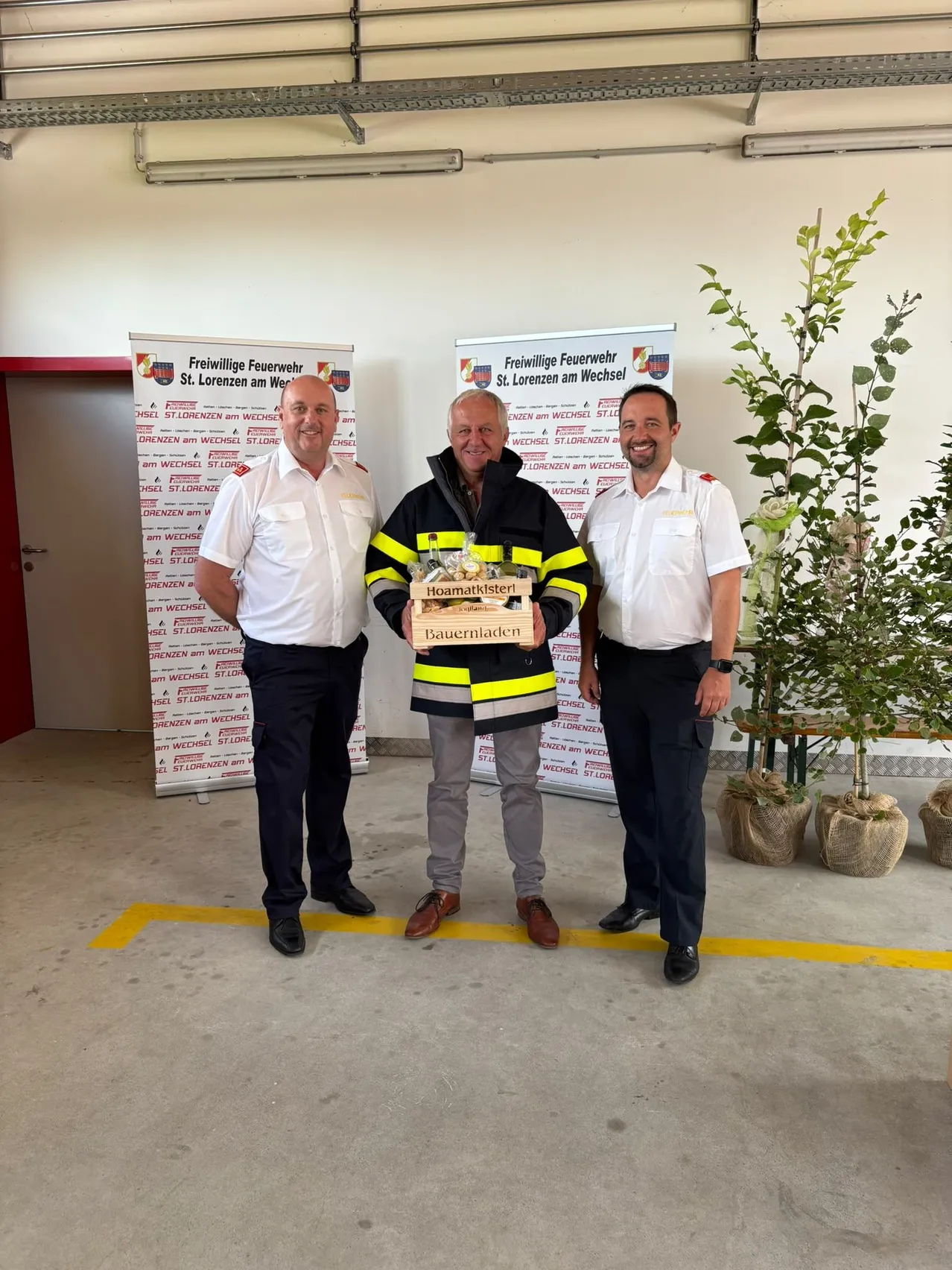 Drei Männer stehen in einem Raum, einer hält eine Box mit der Aufschrift 'Hoamatkläster Bauernladen'. Sie sind in Uniformen gekleidet, der Mann in der Mitte trägt eine Feuerwehrjacke. Hinter ihnen sind Banner mit dem Text 'Freiwillige Feuerwehr St. Lorenzen am Wechsel' angebracht.