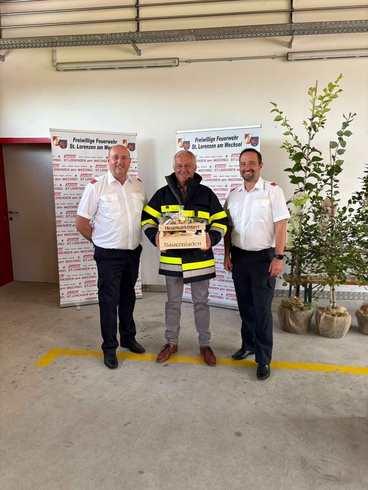 Drei Männer stehen in einem Raum, einer hält eine Box mit der Aufschrift 'Hoamatkläster Bauernladen'. Sie sind in Uniformen gekleidet, der Mann in der Mitte trägt eine Feuerwehrjacke. Hinter ihnen sind Banner mit dem Text 'Freiwillige Feuerwehr St. Lorenzen am Wechsel' angebracht.