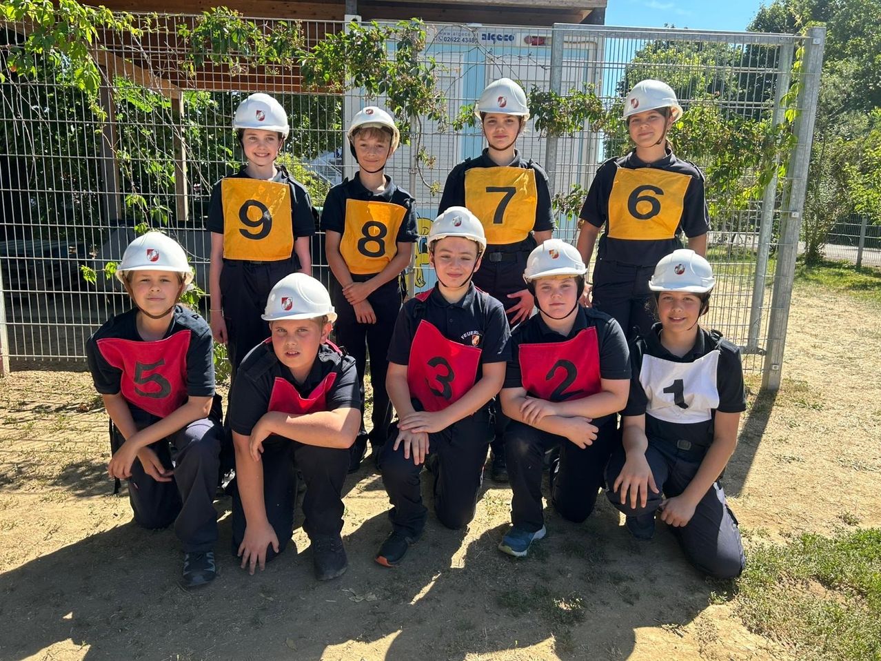 A group of young children, each wearing a white helmet, poses for a photo in an outdoor area with a metal fence and plants. They are wearing numbered vests and seem to be participating in an event.