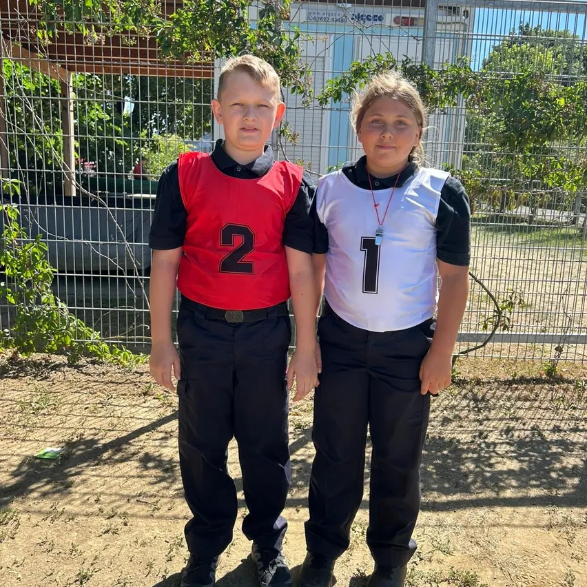 Zwei Kinder in Sportuniformen stehen nebeneinander vor einem Zaun. Der Junge trägt ein rotes Trikot mit der Nummer 2 und das Mädchen ein weißes Trikot mit der Nummer 1.