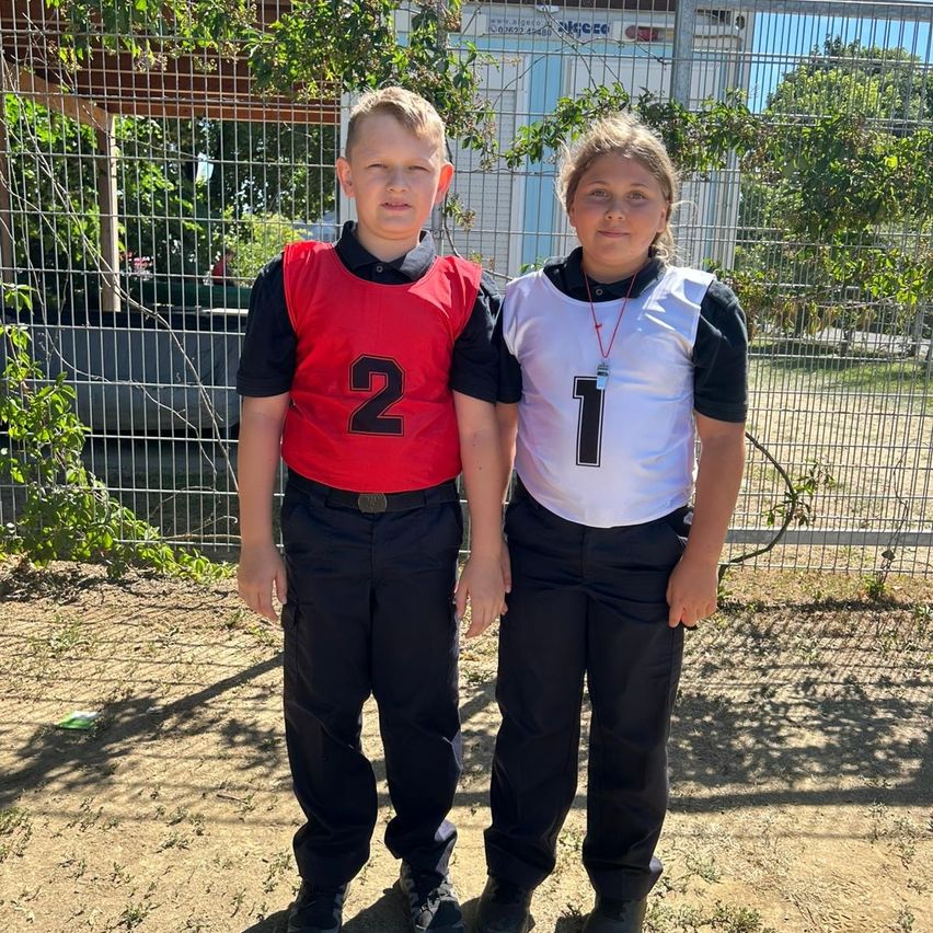 Two children in sports uniforms stand next to each other in front of a fence. The boy wears a red jersey with the number 2 and the girl a white jersey with the number 1.