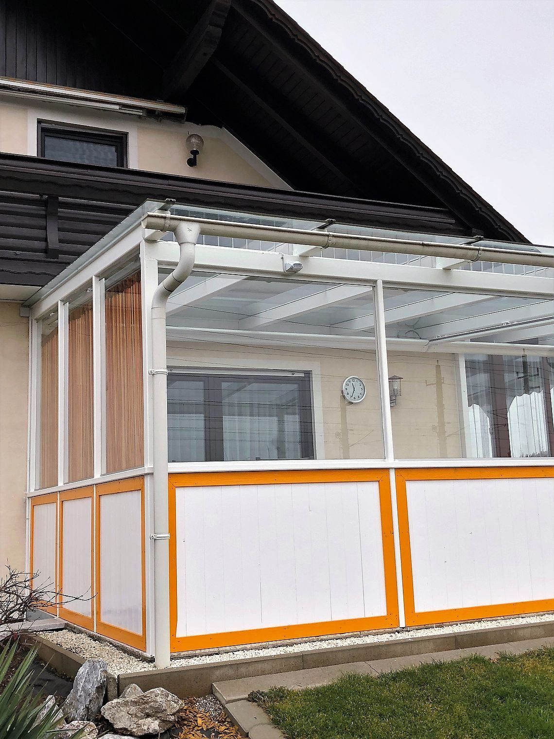 Eine weiße und orange Struktur mit Glasscheiben ist an der Seite eines Hauses befestigt. Es hat ein Glasdach und eine weiße Rohrleitung an der Seite.