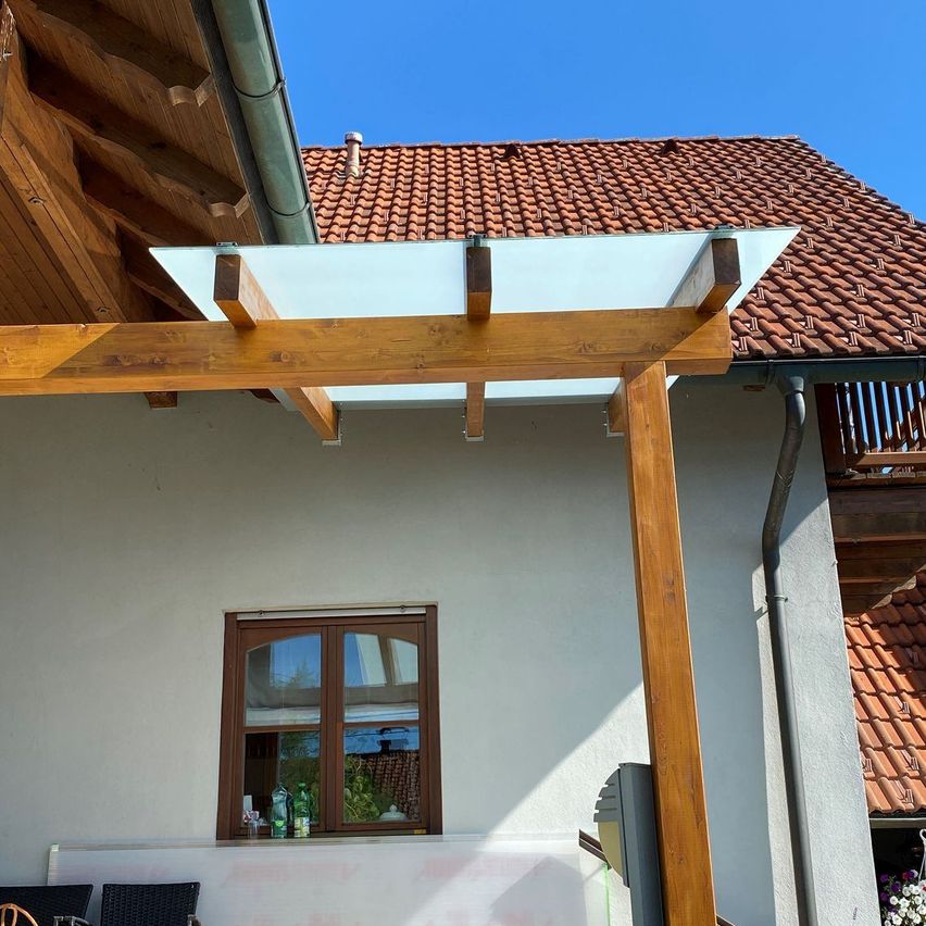 Eine Holzkonstruktion mit einem Glasdach ist an einem Haus mit einem roten Ziegeldach befestigt. Ein Fenster mit einem Holzrahmen spiegelt die Umgebung wider.