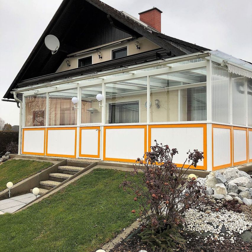 Ein Haus mit einer verglasten Veranda und orange-weißen Akzenten. Die Veranda hat ein Glasdach, einen weißen Vordach und eine Satellitenschüssel auf dem Dach.