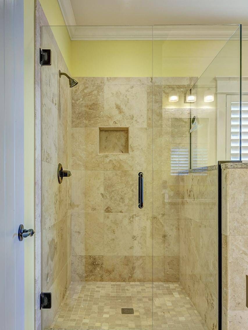 A modern shower area with beige tiles and a glass door, featuring a showerhead and a small shelf.