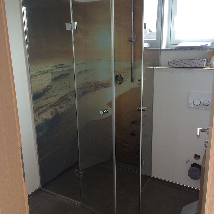 A corner shower with glass walls showing a beach scene. The shower area has a silver handle and a silver faucet.