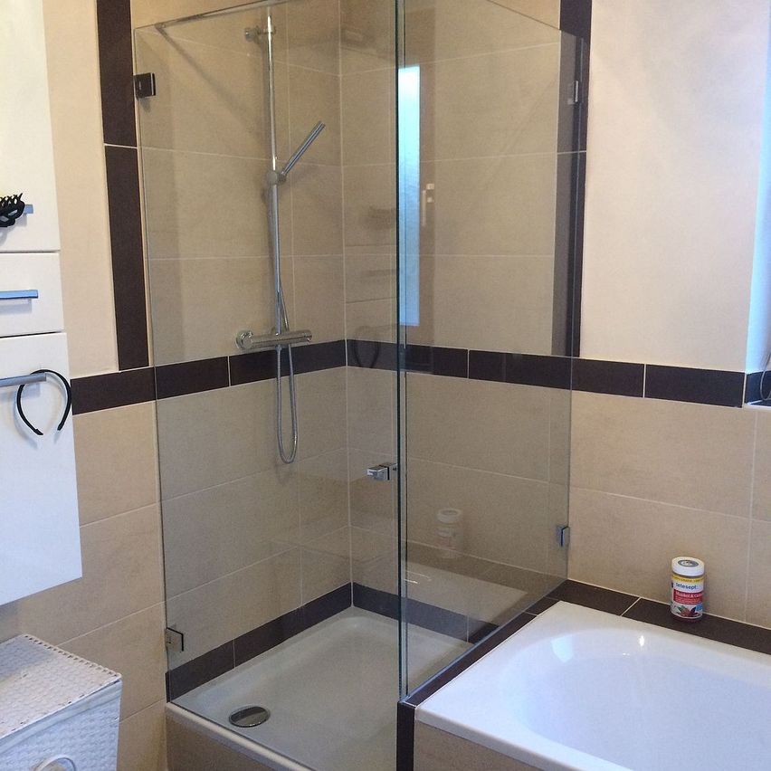 A modern bathroom with a glass shower enclosure, white bathtub, and tiled walls. There is a showerhead and a towel rack.
