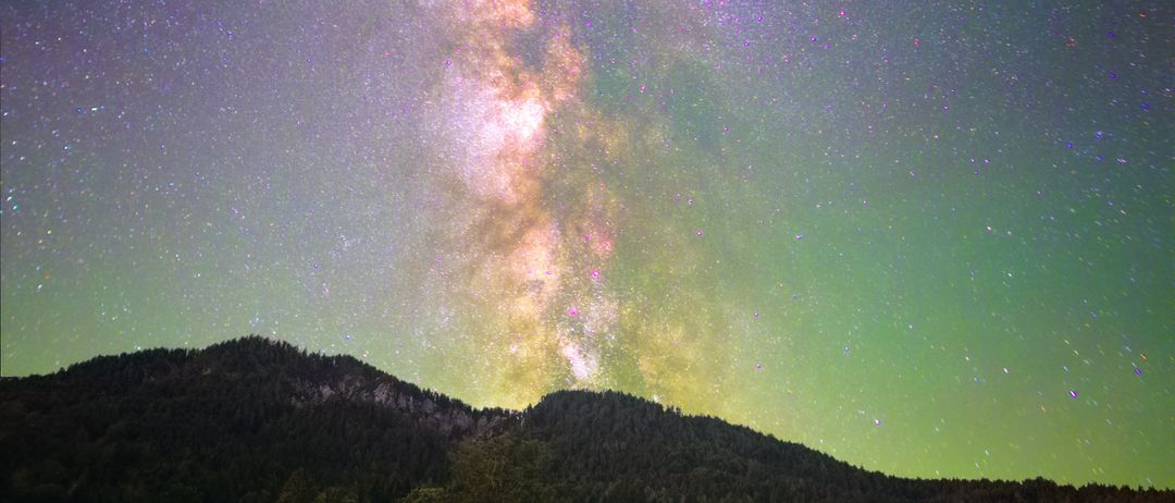 Eine Nachtlandschaft zeigt eine leuchtende Milchstraße, die den Himmel erhellt. Ein Gebirgszug bildet den Hintergrund, und ein kleiner Baum steht in einem grünen Feld. Die Szene wird von einem Kiesweg eingerahmt, der in die Ferne führt.