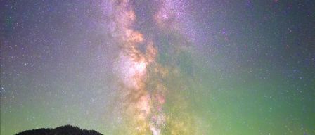 Ein Nachthimmel voller Sterne und der Milchstraße, ein grüner Schimmer in der unteren rechten Ecke und eine Silhouette von Bergen im Vordergrund.