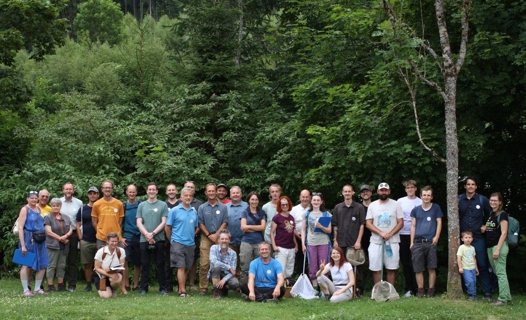 Eine Gruppe von Menschen posiert für ein Foto in einem Wald mit hohen Bäumen und Büschen. Einige stehen, andere sitzen auf dem Gras. Sie alle lächeln und halten Gegenstände wie Bücher, Taschen und Netze.