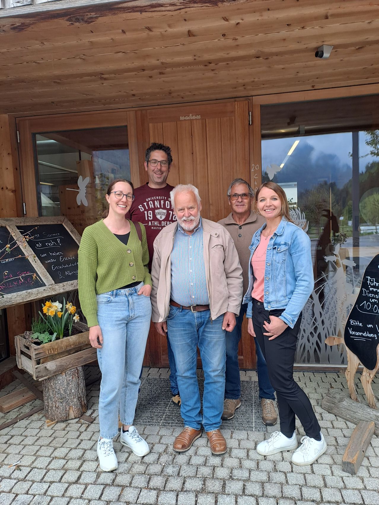 Five people stand outside a wooden building with chalkboards, smiling for a photo. They are surrounded by plants and a wooden table with flowers. The building has glass doors and windows.