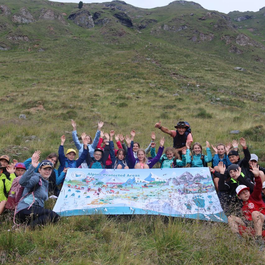 Eine Gruppe von Kindern und Erwachsenen posiert für ein Foto auf einer grasbewachsenen Wiese mit einer großen Karte der Alpenschutzgebiete im Vordergrund.