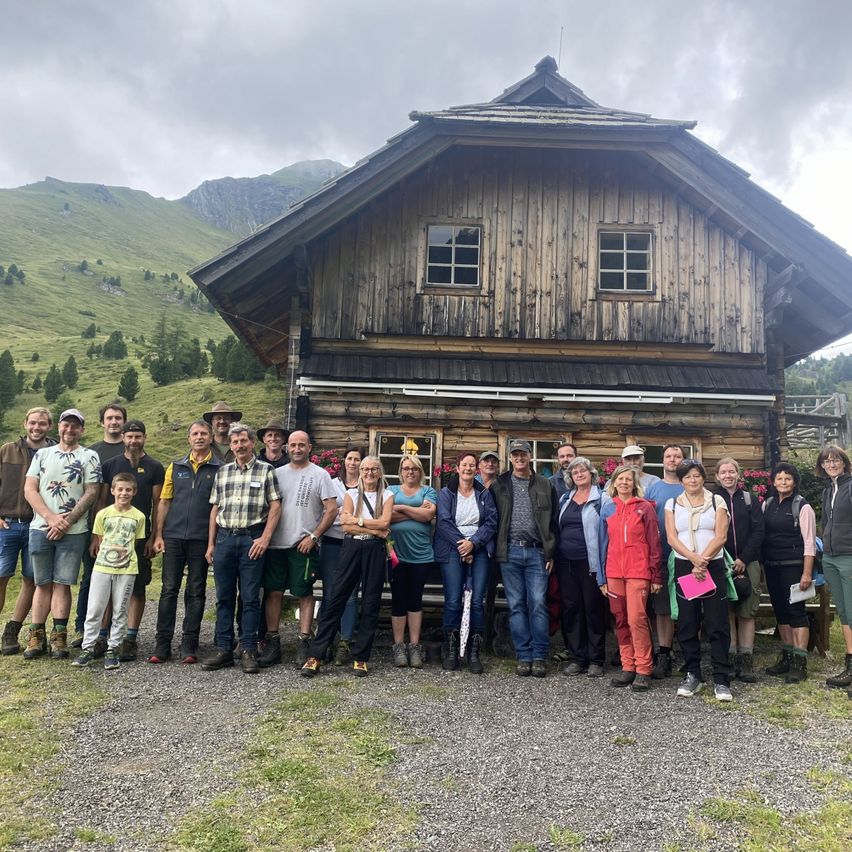 Eine Gruppe von Menschen posiert für ein Foto vor einem Holzhaus mit steilem Dach und einem Berg im Hintergrund.