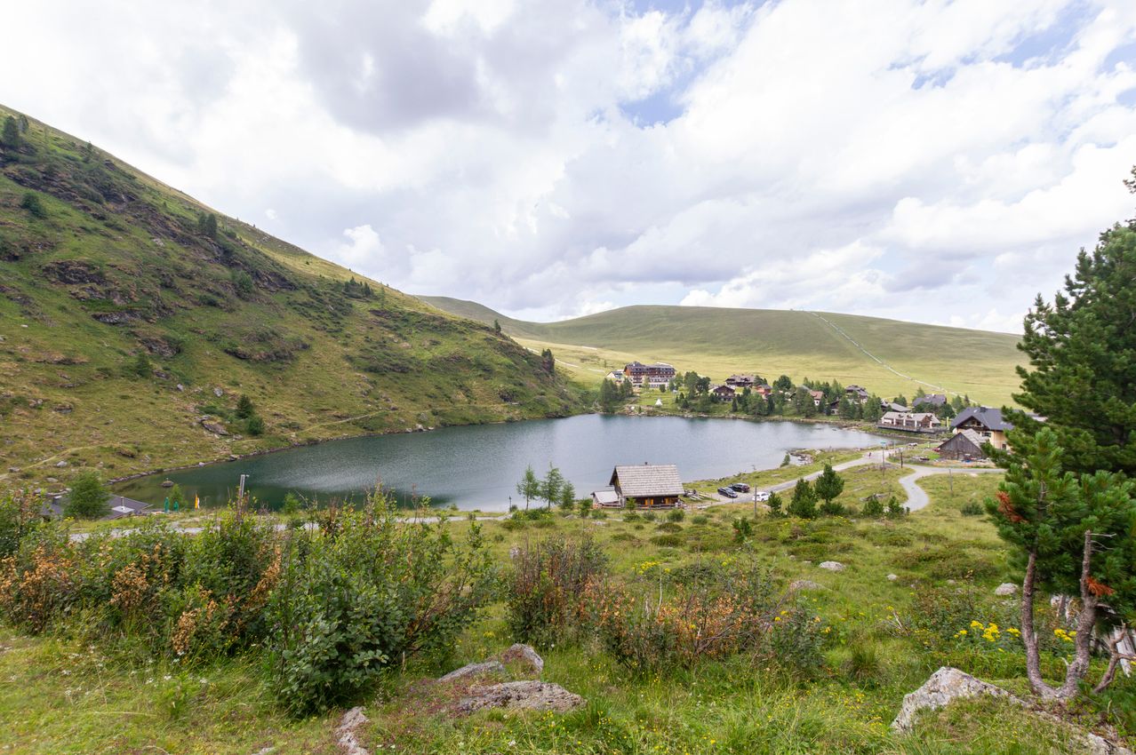 Ein ruhiger Bergsee umgeben von üppigem Grün und einem kleinen Haus am Ufer mit einigen geparkten Autos in der Nähe.