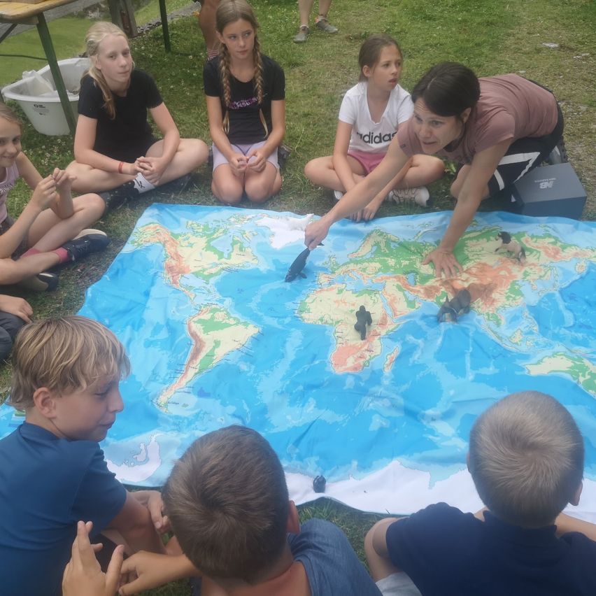 Eine Gruppe von Kindern und einem Erwachsenen sitzen um eine große Weltkarte herum, wobei der Erwachsene auf verschiedene Orte zeigt, während die Kinder aufmerksam zuschauen.