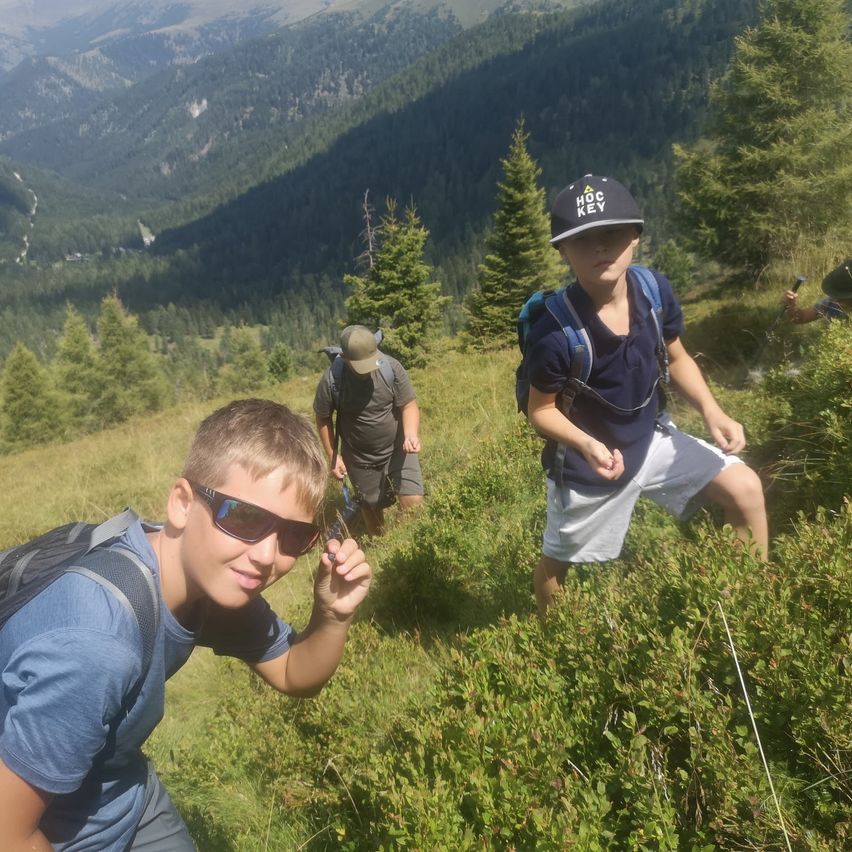 Drei Jungen wandern auf einem grasigen Berghang mit Blick auf ein Tal. Ein Junge trägt eine Sonnenbrille und einen Rucksack.