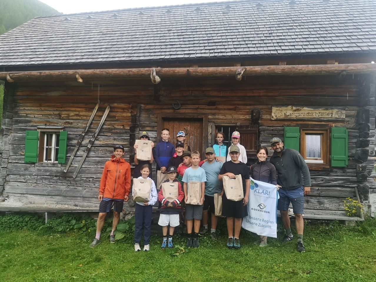 Eine Gruppe von Menschen, darunter Erwachsene und Kinder, posiert für ein Foto vor einem Holzhaus. Sie halten Papiertüten in den Händen, und einige tragen Hüte und Turnschuhe. Ein Banner wird von einer Frau auf der rechten Seite gehalten.