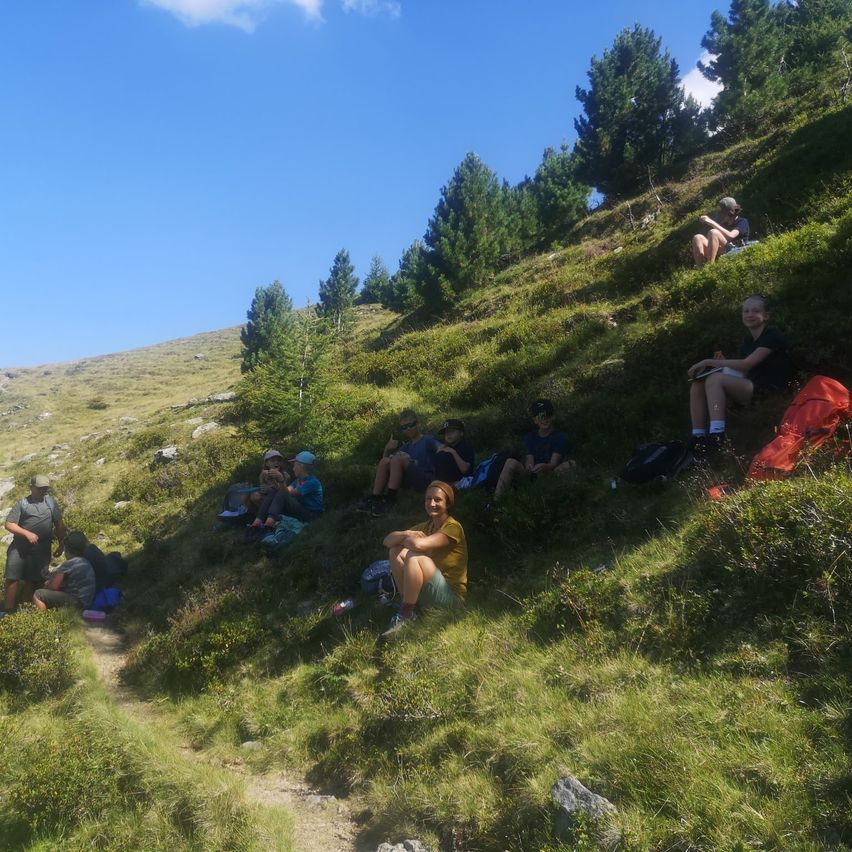 Eine Gruppe Wanderer ruht sich auf einem grasigen Hang unter einem klaren blauen Himmel aus, einige sitzen auf dem Boden und andere lehnen sich gegen den Hang. Rucksäcke und Flaschen sind um sie herum verstreut.