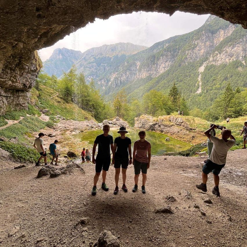 Eine Gruppe von Wanderern steht in einer Höhle mit Blick auf ein Bergtal. Einige machen Fotos, während andere auf einen kleinen Bach zulaufen.