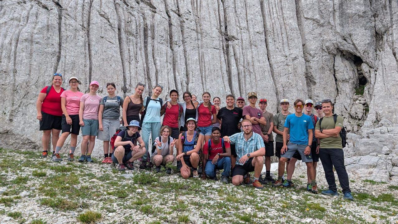Eine Gruppe von Menschen, wahrscheinlich auf einer Wandertour, posiert für ein Foto vor einer großen Felsformation. Einige stehen, während andere knien oder sitzen. Sie alle lächeln und scheinen sich zu amüsieren.