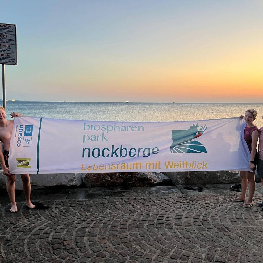 Drei Personen halten ein Banner mit der Aufschrift 'Biosphärenpark Nockberge' mit einem malerischen Blick auf den Ozean im Hintergrund.