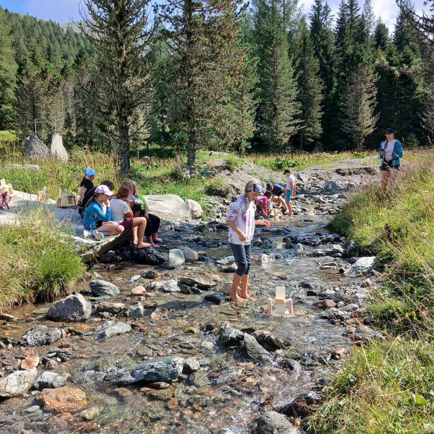 Eine Gruppe von Menschen steht und sitzt in einem Bach, einige tragen Hüte und Brillen. Sie sind von Felsen und Gras umgeben. Bäume sind im Hintergrund.