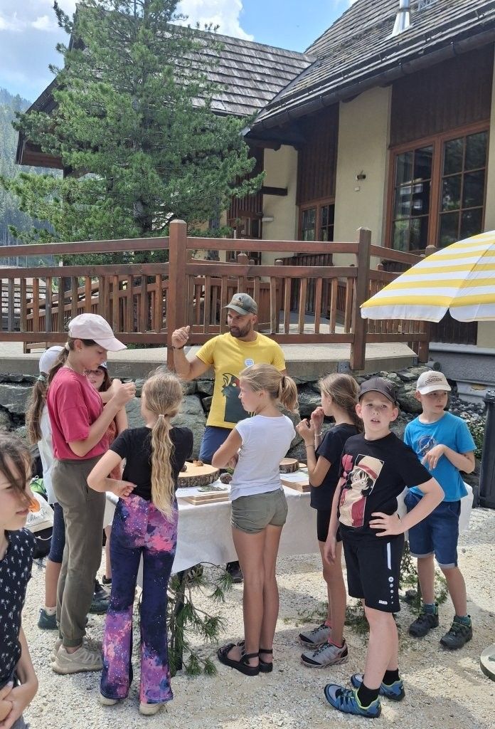 Eine Gruppe von Kindern in Outdoor-Kleidung steht um einen Tisch mit Holz und Messern herum und hört einem Mann in einem gelben Shirt zu.