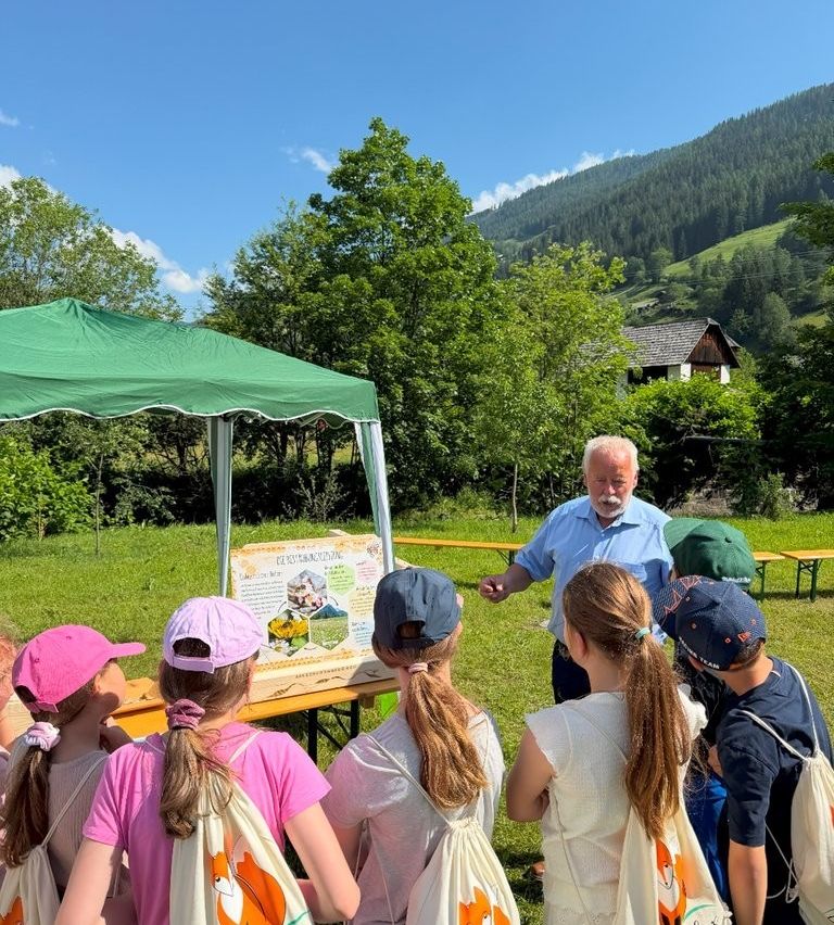 Eine Gruppe von Kindern hört einem Mann unter einem grünen Zelt zu. Der Mann erklärt etwas von einem Brett. Die Kinder tragen Hüte und haben Taschen. Bäume und ein Berg sind im Hintergrund.