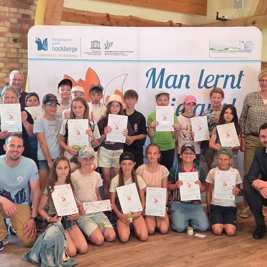 Eine Gruppe von Kindern und Erwachsenen posiert für ein Foto, alle halten Urkunden in den Händen. Hinter ihnen steht ein Banner mit der Aufschrift 'Man lernt gleich'. Sie befinden sich wahrscheinlich bei einer besonderen Veranstaltung.