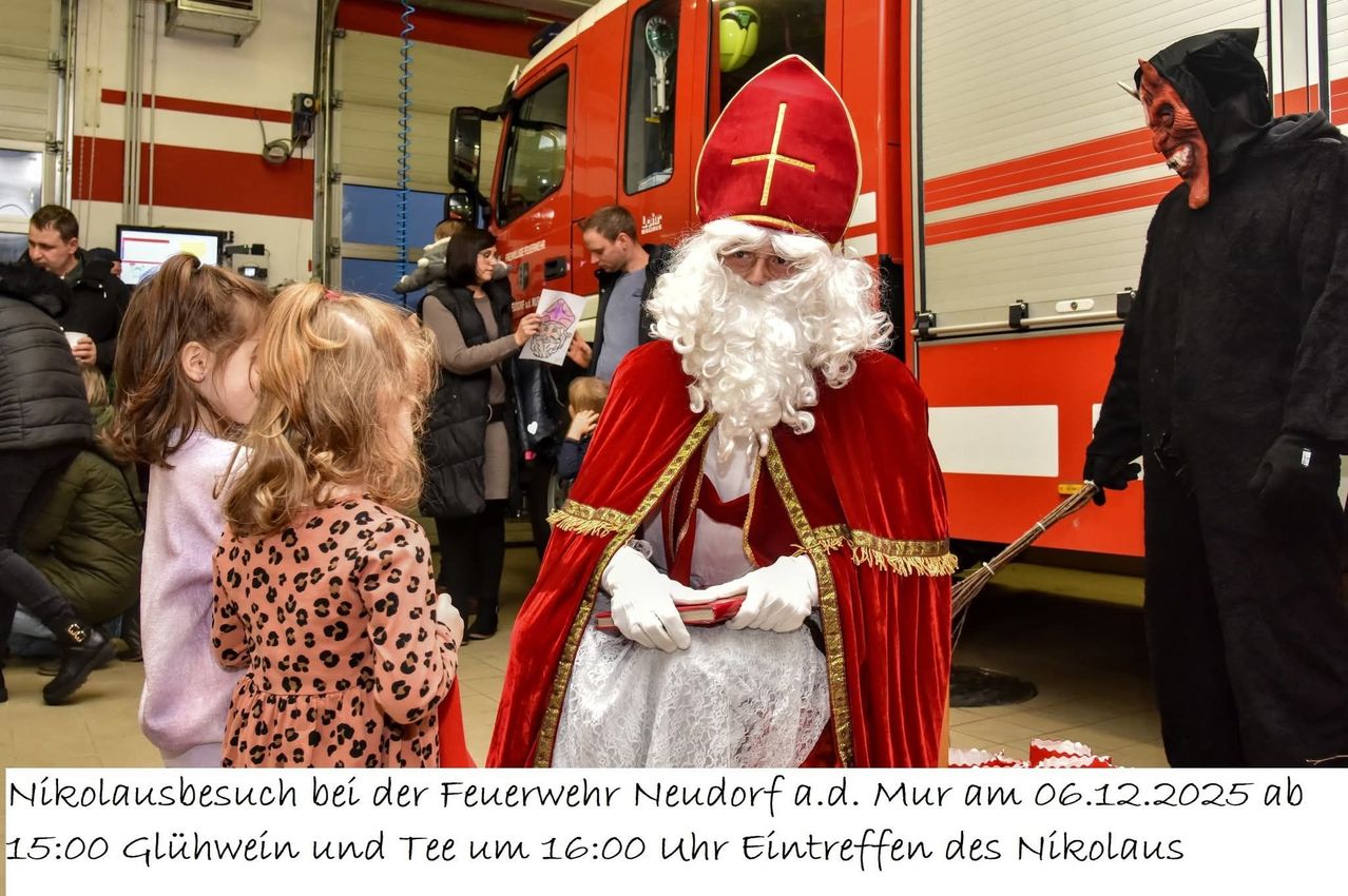 Ein Mann in einer Weihnachtsmann-Verkleidung steht vor einem Feuerwehrauto, hält ein Buch in der Hand, mit Kindern und Erwachsenen in der Nähe. Eine erwachsene Frau hält eine Zeichnung für ihn. Das Bild hat einen deutschen Bilduntertitel.