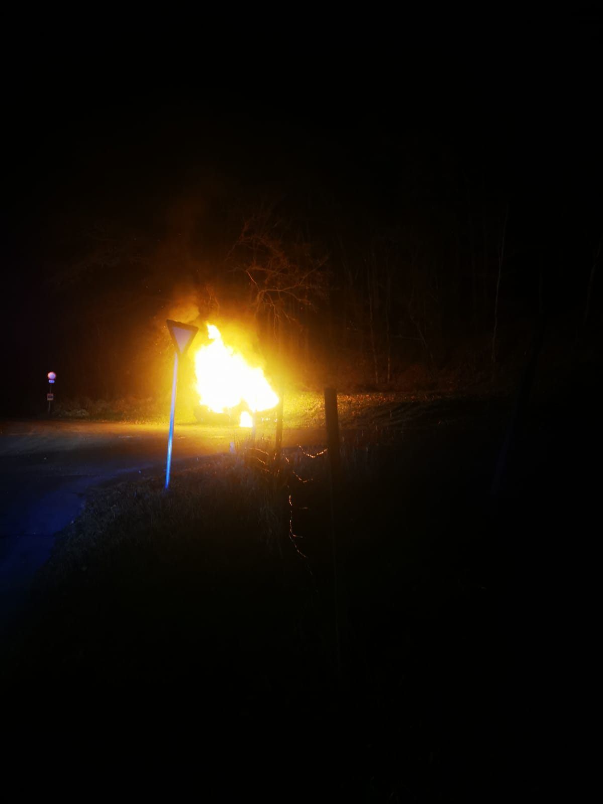 Ein Feuer brennt nachts hell auf einer Landstraße und erhellt die dunkle Umgebung. Ein Pfosten steht in der Nähe und wirft einen Schatten.