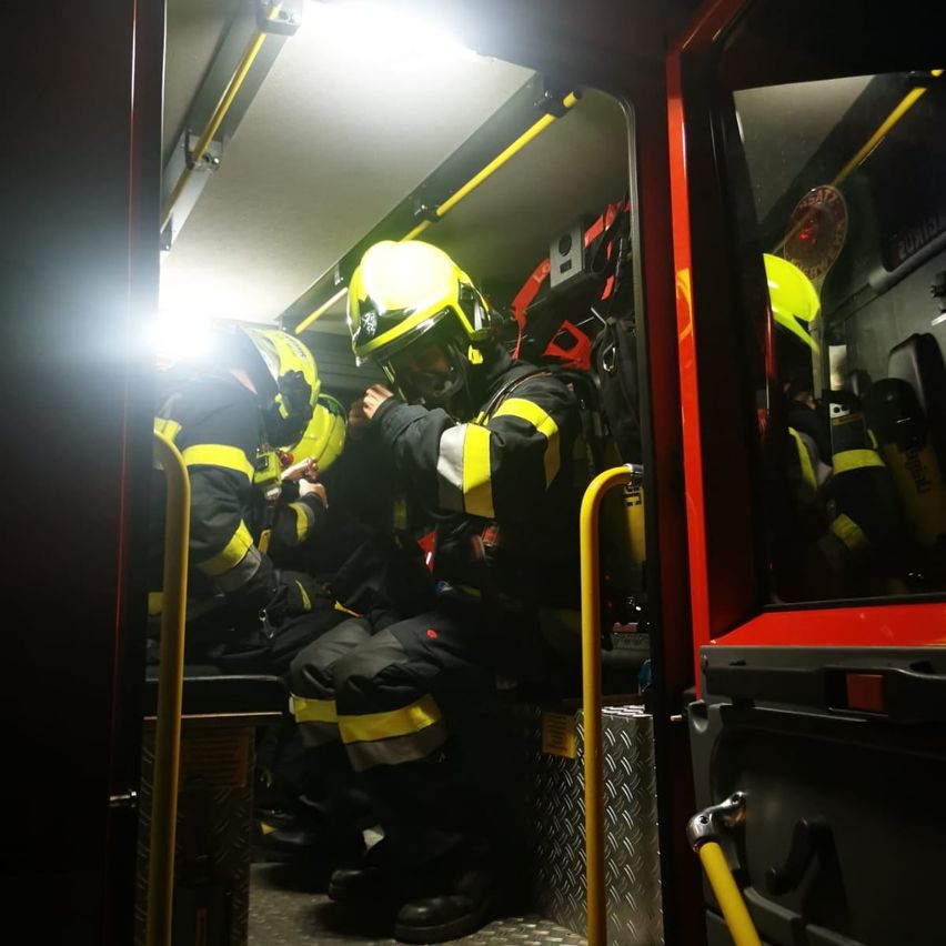 Feuerwehrleute sitzen in einem Fahrzeug, wahrscheinlich einem Feuerwehrauto. Sie tragen gelbe Helme und schwarze und gelbe Uniformen. Ein gelbes Licht beleuchtet das Innere.