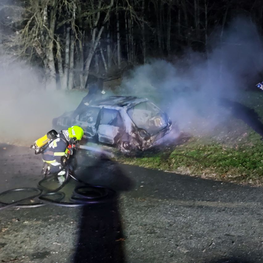 Ein Feuerwehrmann in einem gelben Helm sprüht Wasser auf ein Auto in der Nacht. Das Auto steht am Straßenrand und gibt Rauch ab.