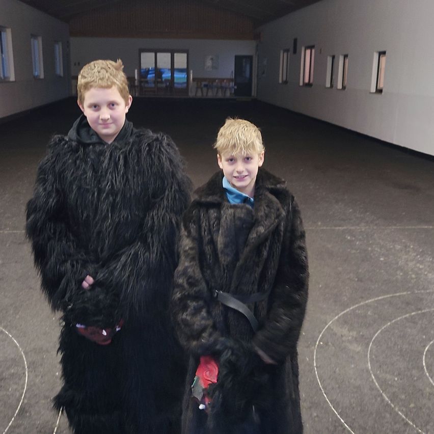 Zwei Jungen in Pelzmänteln stehen lächelnd in einem Flur. Der Junge auf der linken Seite trägt einen roten Handschuh, während der Junge auf der rechten Seite ein rotes Objekt hält. Der Flur ist leer mit sichtbaren Fenstern und Türen.