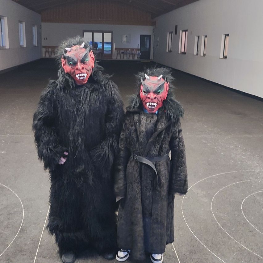 Zwei Personen in pelzigen Kostümen mit gehörnten Masken stehen in einem Raum. Die Person auf der rechten Seite trägt Turnschuhe und einen Gürtel. Hinter ihnen befinden sich leere Tische und Stühle.