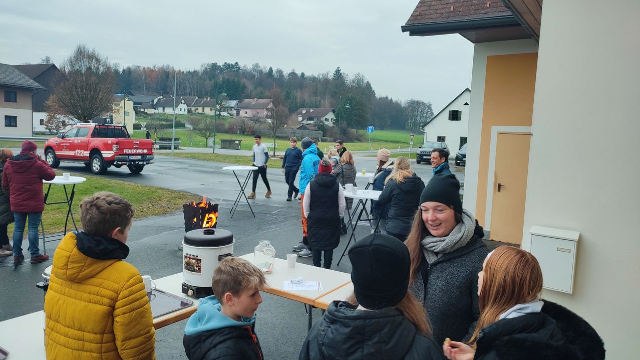 Eine Gruppe von Menschen ist im Freien versammelt. Sie stehen und sitzen um Tische herum, einige mit Tassen. Ein Feuerkorb und ein Reiskocher stehen auf den Tischen. Im Hintergrund befinden sich Häuser und Bäume.