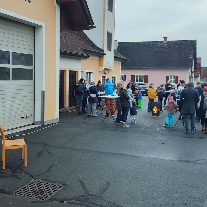 Eine Gruppe von Menschen steht draußen in der Nähe eines Gebäudes, mit einem Stuhl auf der Seite und einem Tisch in der Nähe. Die Umgebung ist draußen, und ein Auto ist im Hintergrund geparkt.